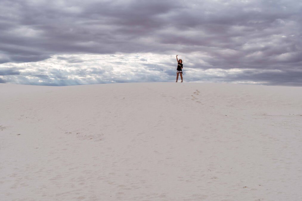 Adult female stands on top of sand dunes in the distance, giving a peace sign at White Sands National Minument